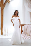 A woman modeling an off-the-shoulder white mermaid-style wedding gown in a bright studio with exposed wooden beams, draped fabric, and soft pink satin drapery.