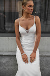 A woman wearing a white spaghetti-strap slip dress with a lace V-neckline, standing against a gray stone wall.