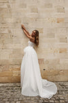 A woman wearing a strapless white wedding gown with a sheer embroidered bodice and a full flowing skirt stands against a beige stone wall on a cobblestone surface.