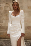 A woman wearing a white long-sleeve evening gown with structured shoulders, a square neckline, and a high slit, standing against a beige stone wall.