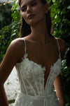 A woman wearing a white lace corset-style dress with thin straps, photographed outdoors against a background of green foliage.