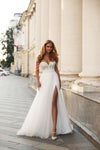 A woman wearing a white wedding gown with a thigh-high slit, standing on a city sidewalk in front of a classical building with large columns.