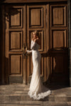 A woman in a fitted off-the-shoulder white lace wedding gown stands in front of large dark wooden doors, holding one of the handles. The dress has long sleeves, a fitted silhouette, and a slight train at the hem.