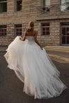 A bride wearing a strapless white wedding gown with a structured corset-style bodice and voluminous tulle skirt, photographed from behind while holding out the train in front of a stone building.
