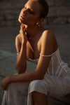 A woman seated outdoors in warm sunlight wearing a sheer white lace dress with pearl-like off-the-shoulder straps and matching pearl earrings.