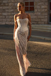 A woman wearing a strapless corset-style maxi dress with a sheer, textured skirt walks outdoors on a paved street in front of a stone building.