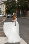 A woman wearing a voluminous off-the-shoulder white wedding gown stands on a city street, holding out the train of the dress. The background shows blurred buildings, trees, and parked cars.