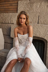 A woman wearing a strapless white lace wedding dress with long sheer lace sleeves sits against a stone wall. The dress features a structured lace bodice with layered tulle and a full skirt.