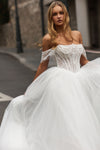 A woman wearing an off-the-shoulder white lace wedding gown with a structured corset bodice and a voluminous tulle skirt, photographed outdoors on a street.