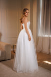 A woman wearing a strapless white wedding gown with a fitted bodice and full tulle skirt, standing in a softly lit indoor room with curtains and a sofa in the background.