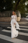 A woman wearing a white lace wedding dress stands on a city crosswalk with trees and a fence in the background.