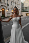A woman wearing an elegant off-the-shoulder white gown with a structured corset-style bodice and flowing skirt, standing on a city street with buildings in the background.