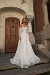 A woman wearing a white off-the-shoulder lace wedding gown with long sheer sleeves and a full skirt, standing on stone steps in front of a building with a wooden door.