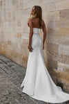 A woman wearing a strapless white wedding gown stands with her back to the camera against a beige stone wall on a cobblestone street.