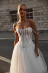 A woman wearing a strapless white wedding gown with a structured corset-style bodice and full tulle skirt, standing outdoors in front of a stone building.