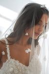 A close-up of a bride wearing a white floral lace wedding dress and a sheer white veil decorated with small white flower appliqués. Her dark hair is styled with matching white flower clips, and she is wearing white flower earrings.