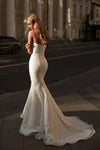 A woman wearing a strapless white lace mermaid wedding gown stands on a city sidewalk with classical columns and a street in the background, captured in warm golden-hour sunlight.