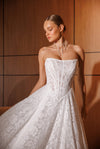 A woman wearing a strapless white lace wedding gown with a fitted bodice and full skirt, posed against a warm wood-paneled background.