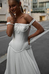 A woman wearing a white off-the-shoulder A-line dress with pearl-like beaded detailing on the bodice, standing on a city street.