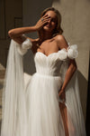 A woman modeling a white off-the-shoulder wedding gown with voluminous tulle sleeves, a sweetheart neckline, and a long sheer train against a warm-toned indoor wall.