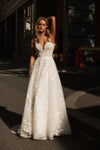 A woman wearing a strapless white A-line wedding gown with sparkling sequin embellishments, standing outdoors on a street in dramatic sunlight and shadow.