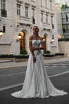 A woman modeling an elegant off-the-shoulder A-line wedding gown with a fitted bodice and flowing skirt, standing on a city street in front of a grand classical building.