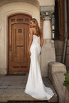 A woman wearing a strapless white mermaid-style wedding gown stands on stone steps in front of a wooden door and stone column, photographed from the back with the train of the dress visible.
