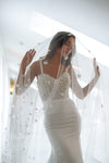 A woman wearing a white lace wedding dress and a long sheer veil with small floral embellishments, posing indoors with her hands holding the veil out to the sides.