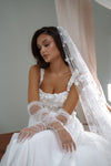 A woman wearing a white lace bridal gown with sheer tulle opera gloves and a long floral lace veil, seated indoors against a soft neutral background.