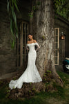 A woman wearing an off-the-shoulder white lace wedding gown with a fitted bodice and flared mermaid skirt, standing beside a large tree outdoors in front of a stone building.