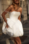 A woman wearing a strapless white mini dress with a structured corset-style bodice, dramatic voluminous gathered skirt, and pearl-like chain embellishments across the shoulders, photographed outdoors on a city street.