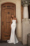 A woman wearing a strapless white mermaid-style wedding gown stands in front of a large wooden door and stone column.