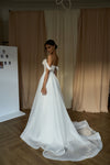 A woman wearing a white off-the-shoulder wedding gown with a long flowing train, standing in an indoor room with beige curtains and wood flooring.