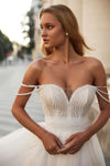 A woman wearing a strapless white wedding gown with a heavily beaded sweetheart bodice and pearl-like shoulder straps, photographed outdoors with a softly blurred background.