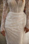 Close-up of a white lace wedding dress with long sheer sleeves, a deep V-neckline, and a ruched gathered waist, shown on a model.