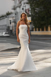 A woman stands on a city street wearing a strapless white mermaid-style gown with a fitted bodice and flared skirt. The background shows blurred buildings, trees, and pavement.