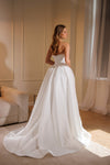 A woman wearing a strapless white wedding gown with a fitted bodice and a full flowing skirt, photographed indoors in a softly lit room with curtains and a sofa in the background.
