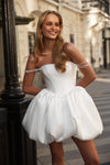 A woman modeling a strapless white puff-skirt dress with pearl chain accents on the off-the-shoulder straps, standing on a city street.