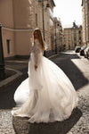 A woman wearing a voluminous white wedding gown stands on a sunlit city street, with the dress spread wide around her and buildings lining the background.
