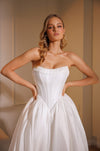 A woman modeling a strapless white wedding gown with a structured corset-style bodice and a full flowing skirt, posed indoors against a warm beige wall.