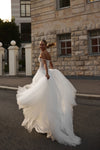 A woman wearing a voluminous white wedding gown with a strapless bodice and full tulle skirt, photographed outdoors on a street in front of a stone building.