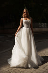 A woman wearing a white wedding gown stands outdoors on a paved street at sunset. The dress has a fitted, beaded strapless bodice and a voluminous, flowing tulle skirt.
