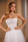 A woman modeling a strapless white wedding dress with a structured lace corset bodice and a full tulle skirt, posed against a softly lit beige interior background.