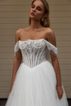 A woman wearing a white off-the-shoulder wedding dress with a lace corset bodice and a full tulle skirt, standing against a beige paneled wall.