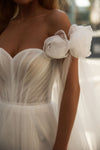 Close-up of a white off-the-shoulder wedding dress with a sweetheart neckline, pleated bodice, and large ruffled fabric flowers on the shoulders.