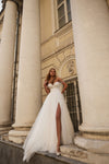 A woman wearing a white wedding gown with a thigh-high slit, standing on stone steps between large classical columns in front of a building facade.
