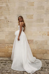 A woman wearing a white strapless wedding gown with a structured bodice and full flowing skirt, standing against a beige stone wall on cobblestone ground.
