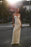 A woman wearing a floor-length ivory wedding gown stands on an outdoor city street at sunset. The dress has thin straps, a fitted bodice, and a deep V-neckline with subtle embellishment.
