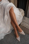 A close-up of a person seated on stone steps wearing a white wedding dress and pointed lace shoes.