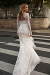 A woman wearing a white lace wedding dress with long sheer sleeves and a fitted mermaid-style skirt, photographed outdoors on a street crosswalk.